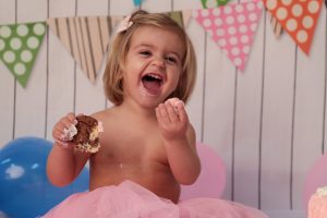 niña riendo mientras sostiene en una mano un trozo de galleta y en la otra tarta en una sesion smash cake